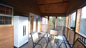 Un porche abrité avec une terrasse en bois, une table en verre, des chaises en métal, un réfrigérateur blanc et une porte donnant sur l’extérieur.