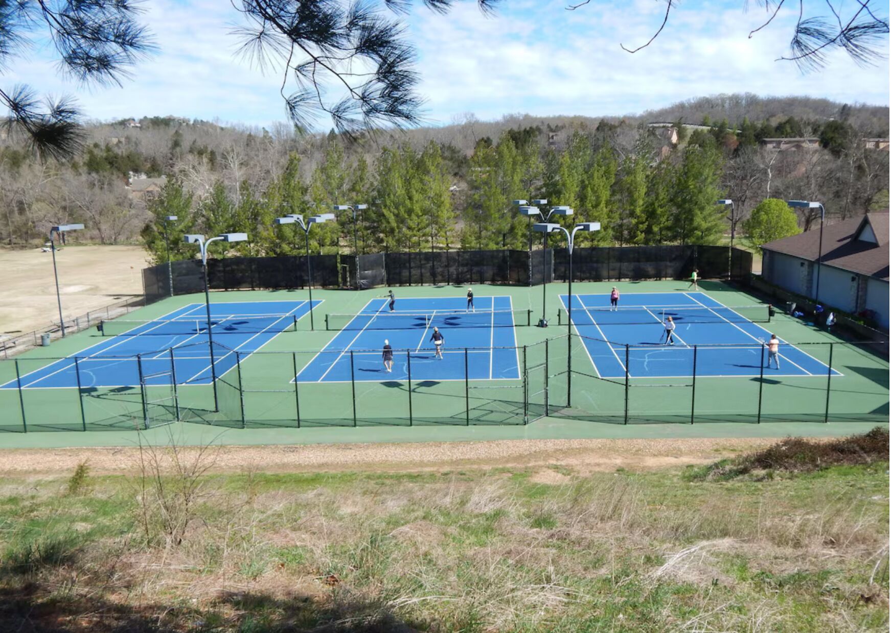 Complejo de canchas de tenis al aire libre con múltiples canchas, rodeado de árboles y un edificio al fondo.