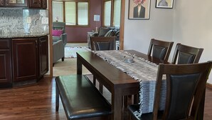 A dining area with a wooden table, chairs, and benches. There is a kitchen area with cabinets and a chandelier.