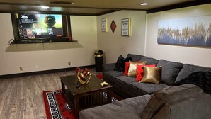 A modern living room with a grey sofa, red and gold cushions, a wooden coffee table, and a red and black patterned rug. A flat-screen TV is mounted on the wall, and there are two framed motivational quotes.