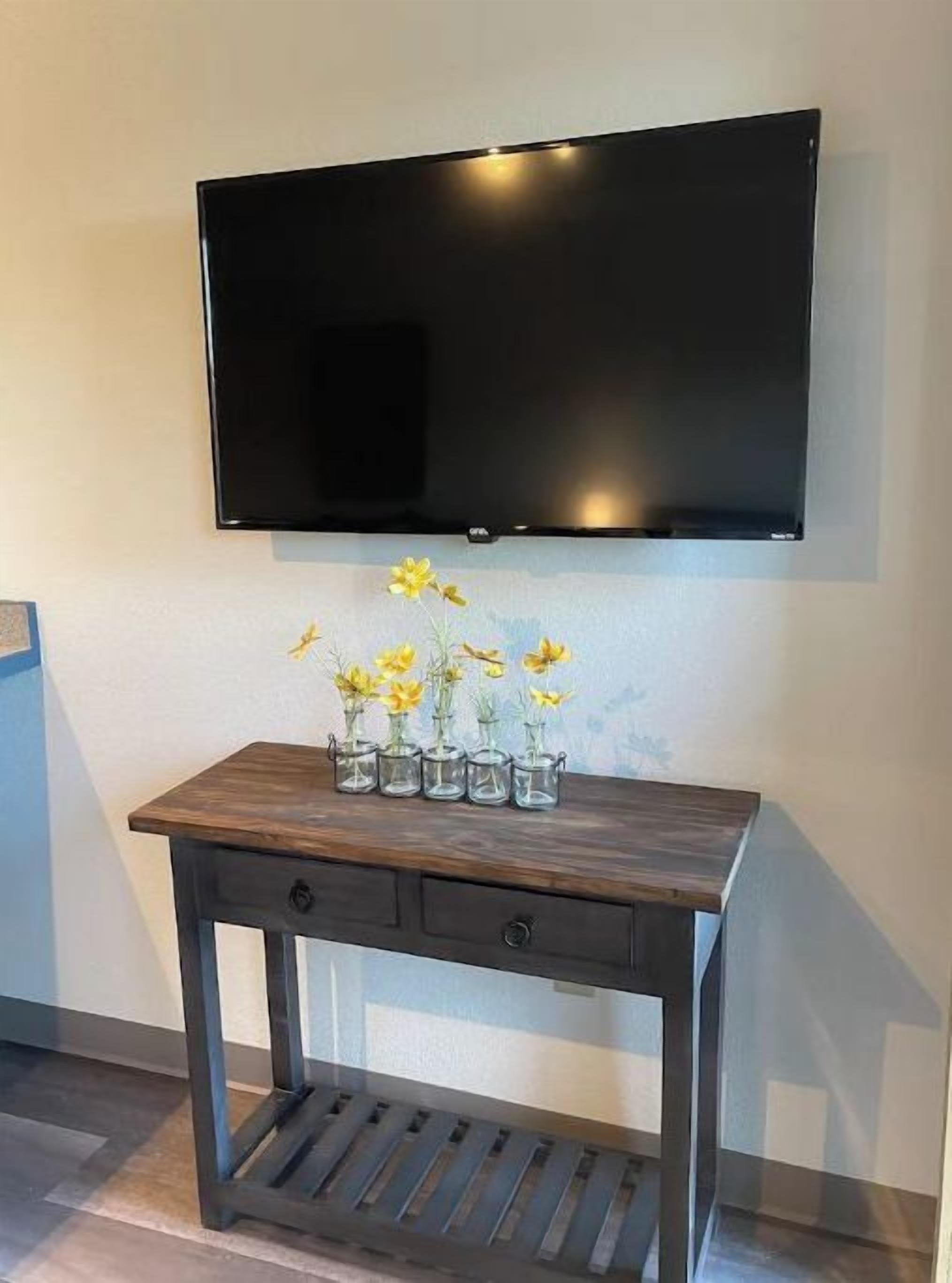 Un téléviseur à écran plat fixé au mur, au-dessus d’une console en bois sur laquelle est posé un vase rempli de fleurs jaunes.
