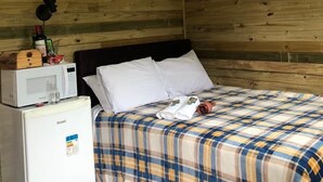 Een kleine, gezellige kamer met een houten plafond en muren, een bed met een geruite deken, een witte koelkast en een magnetron erboven.