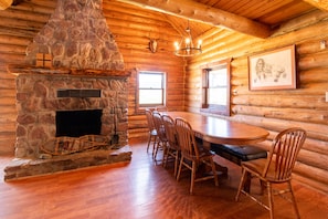 A log cabin interior with a stone fireplace, wooden dining table, and chairs. - East Glacier Cabin w/ Stunning Views of the Rockies! 30 min from 2 GNP entrances (Browning)