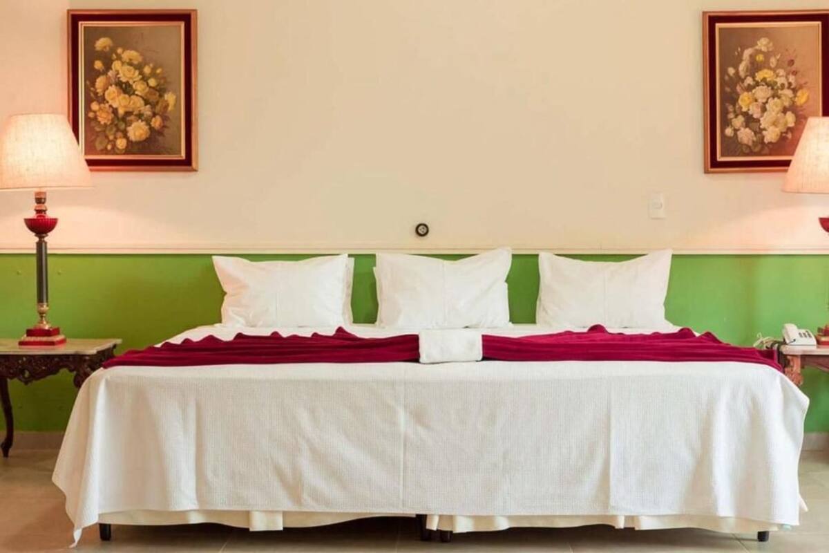 Un lit bien fait, avec des draps blancs et une couverture rouge, encadré de têtes de lit peintes en vert et agrémenté de deux tableaux floraux encadrés accrochés aux murs.