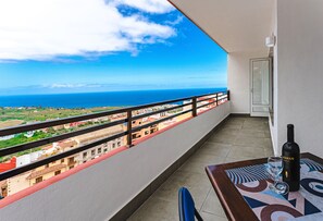A balcony with a view of the ocean and buildings, a table with a bottle and glass, and a door.