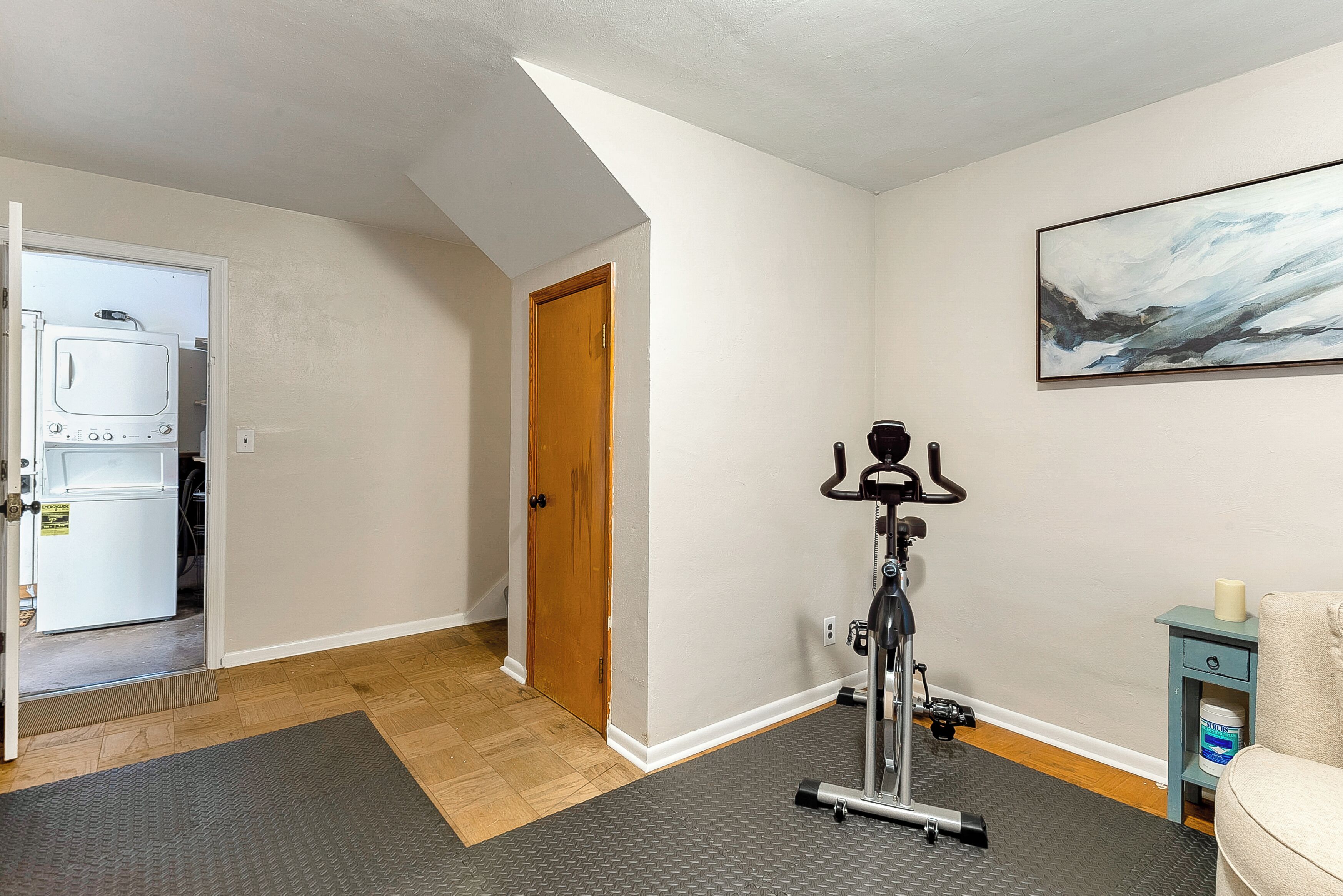 A compact room with a door leading to a laundry area, a standing exercise bike, and a small table with a container.
