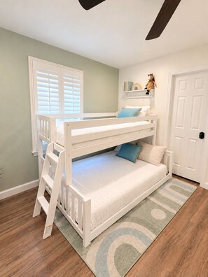 A bunk bed with a ladder, a window with blinds, a door, and a shelf with books and a stuffed toy.