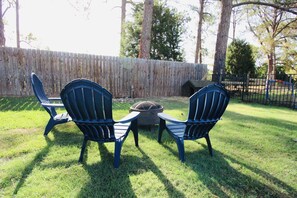 Two outdoor chairs facing a fire pit in a grassy yard with a wooden fence and trees in the background.