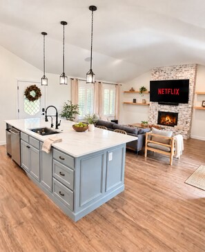 A modern kitchen with a central island, pendant lights, a fireplace, and a flat-screen TV mounted above it.