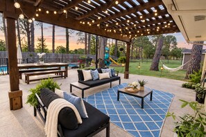 An outdoor patio with a wooden pergola, string lights, a pool, and a children's play area.