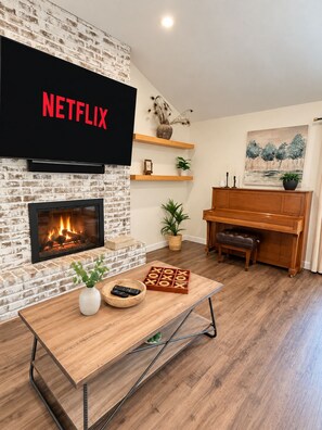 A living room with a fireplace, a television mounted on a brick wall, a wooden coffee table, a piano, and a plant.