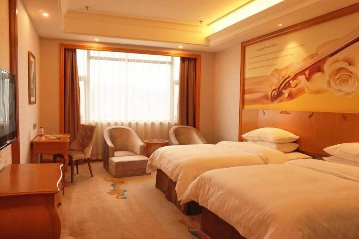 A hotel room with two beds, a desk, a chair, a TV, and a large window with curtains.