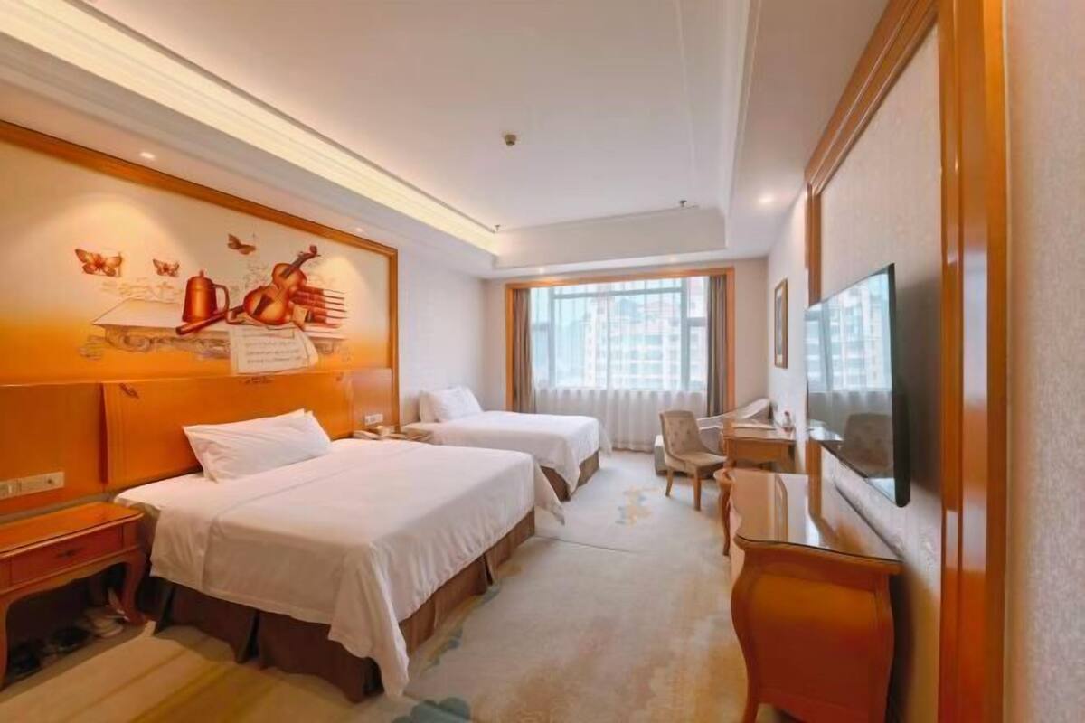 A hotel room with two beds, a desk, a chair, a TV, and a window.