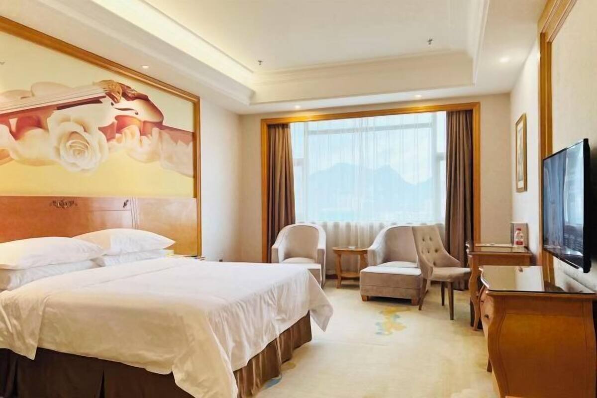 A hotel room with a large bed, two armchairs, a small table, a TV, and a painting on the wall.