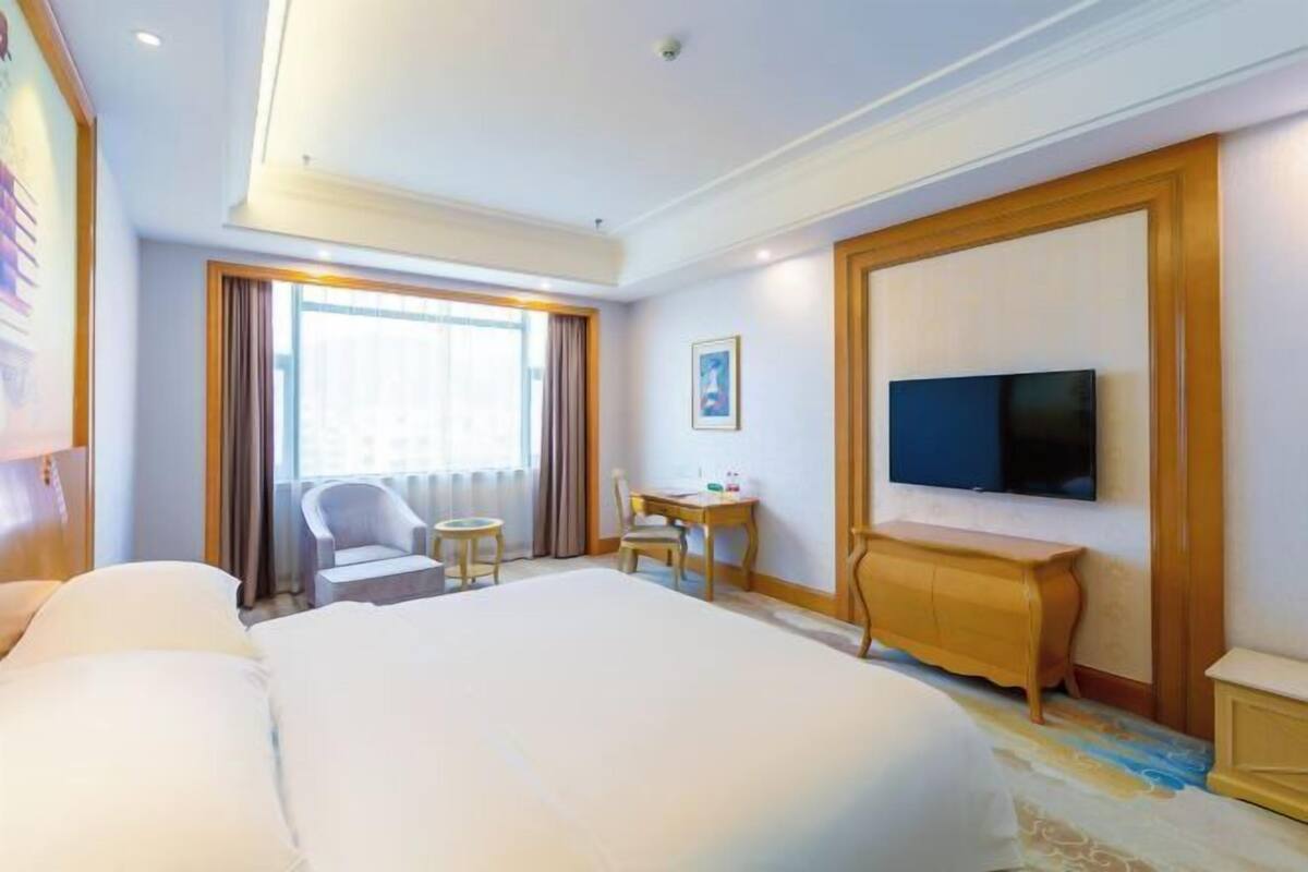 A hotel room with a large bed, a desk with a chair, a television, and a window with curtains.