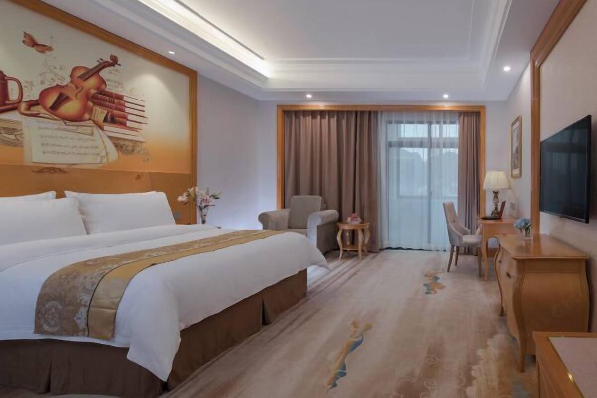 A hotel room with a large bed, a desk, a chair, a TV, and a window with curtains.