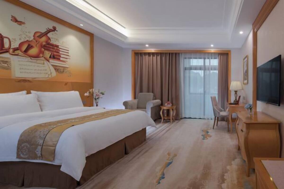 A hotel room with a large bed, a desk, a chair, a TV, and a window with curtains.