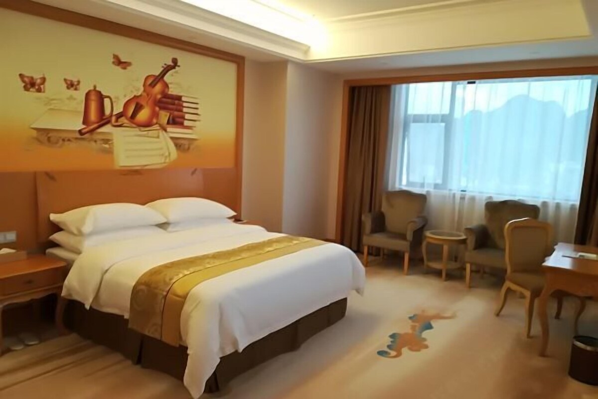 A hotel room with a large bed, a desk, a chair, and a window with curtains.