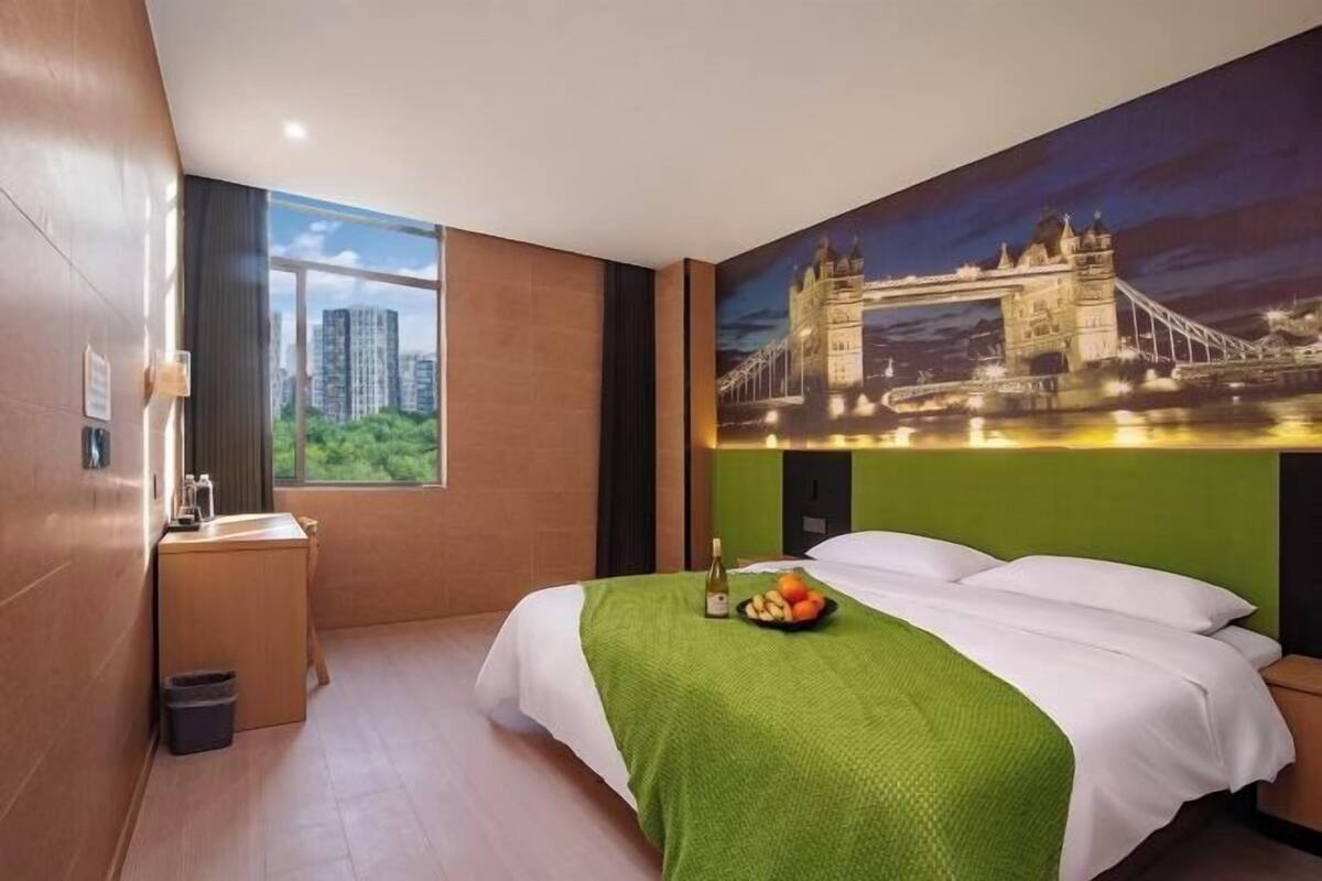 A hotel room with a bed, a desk, a trash bin, a bottle of wine, and a bowl of fruit.