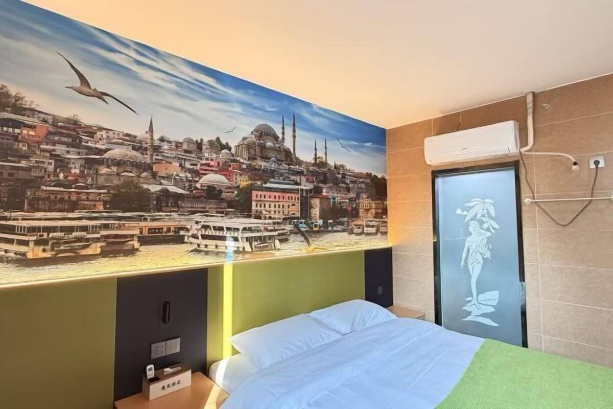 A hotel room with a bed, a wall mural of a cityscape, a window with a view of the city, and a towel rack.