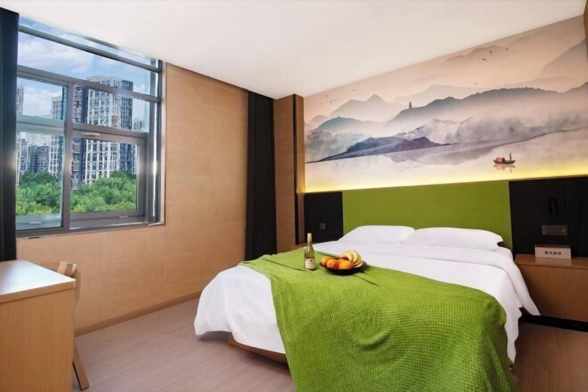 A hotel room with a bed, a desk, a window with a view of buildings, and a mural on the wall.