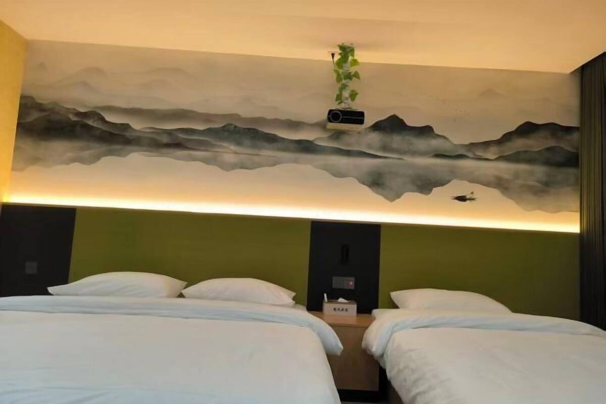 A hotel room with two beds, a wall mural, a plant, and a nightstand with a lamp.