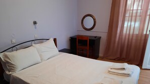 Una cama bien tendida con sábanas y toallas blancas, un espejo redondo en la pared, una silla de madera y un escritorio con una computadora.