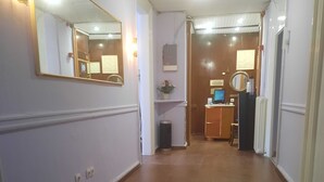 Un pasillo con suelo de madera, un espejo grande a la izquierda y una puerta al final que da a una habitación con un escritorio y una computadora.