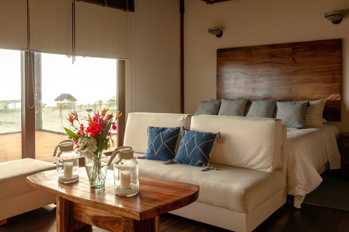 A bedroom with a large bed, a sofa, a wooden table with candles, and a view of the beach.