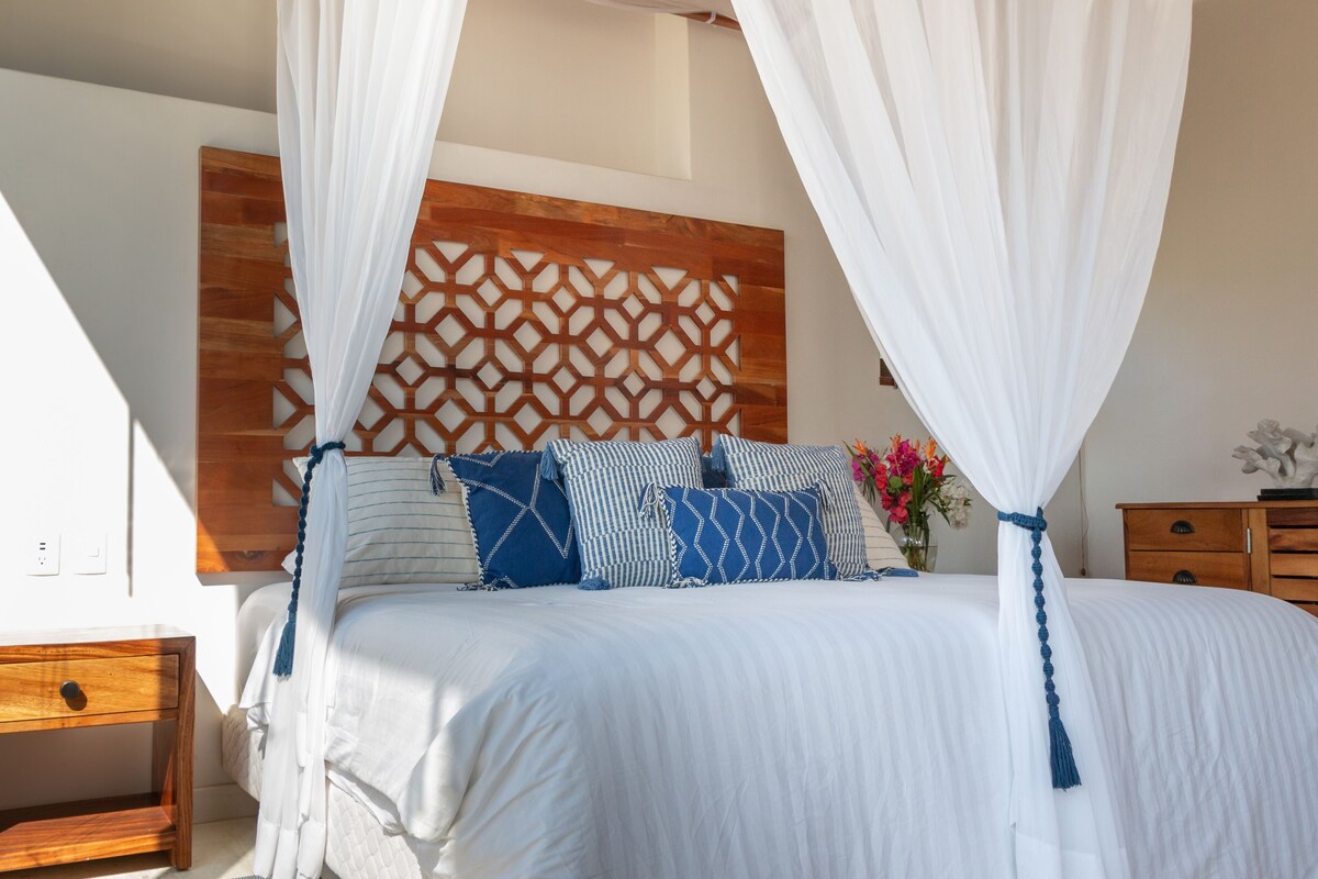 A bedroom with a bed, wooden headboard, white curtains, and a nightstand.