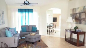 Ein Wohnzimmer mit grauem Sofa, Hocker und Couchtisch. Es gibt einen Essbereich mit Tisch und Stühlen sowie einen an der Wand befestigten Fernseher.