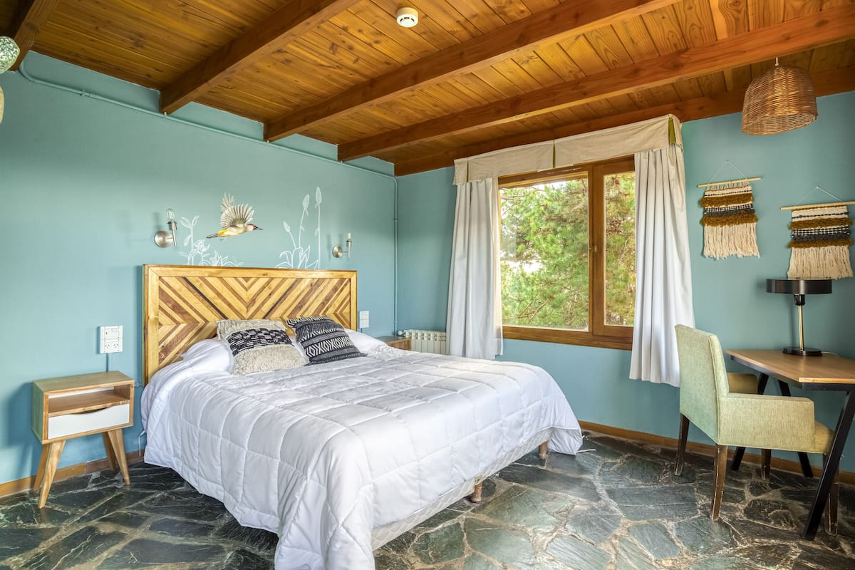 A bedroom with a large bed, a wooden headboard, a stone floor, a window with curtains, and a small table with a lamp.