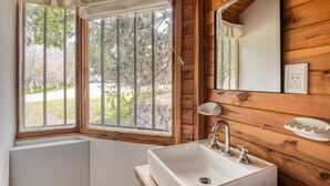 Une salle de bain avec un lavabo blanc, des murs en bois, une fenêtre donnant sur des arbres et un miroir.