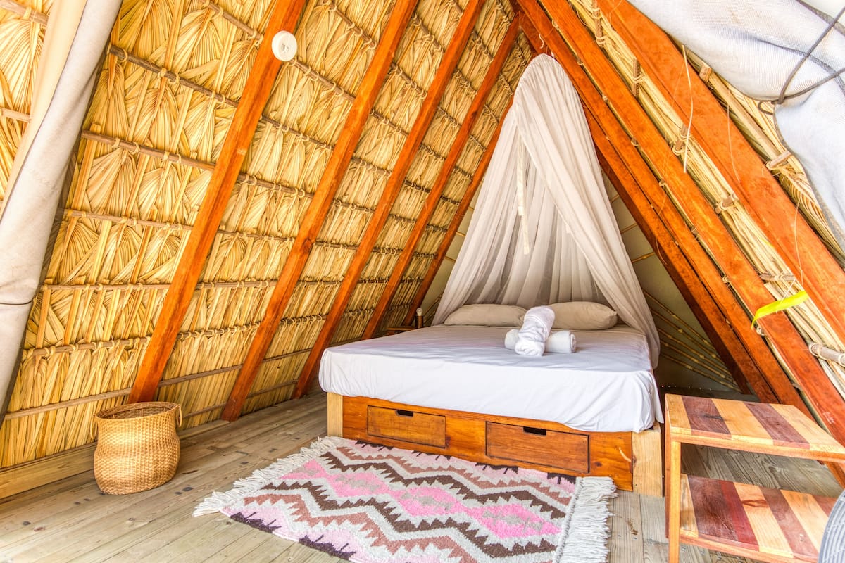 A cozy, triangular room with a thatched roof, a bed with a canopy, and a woven basket.