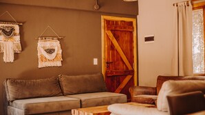 Un salon chaleureux avec un canapé marron, une table basse en bois et un fauteuil en cuir. On peut également apercevoir une porte en bois et une décoration murale en macramé.