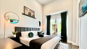 A modern bedroom with a large bed, a bedside table, a mirror, a wall-mounted TV, and a window with curtains. - Modern & Bright Townhouse 26th Ave (Nashville)