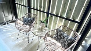 A balcony with two wicker chairs and a small table, a potted plant, and a view of a building with vertical wooden slats. - Modern & Bright Townhouse 26th Ave (Nashville)