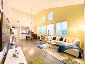 A spacious living room with a large sofa, a dining area with a table and chairs, and a kitchen in the background. - High Ceiling & Amazing backyard & family Kids friendly (Elk Grove)