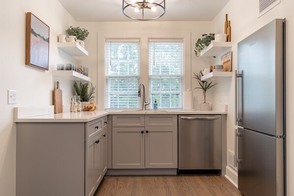 A modern kitchen with grey cabinets, a stainless steel refrigerator, and a window with blinds. - 51 Church Street, Lenox MA - One Bedroom Unit (Lenox)