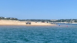 Une plage avec une eau d’un bleu limpide, un rivage sablonneux, et quelques personnes et véhicules visibles au loin.