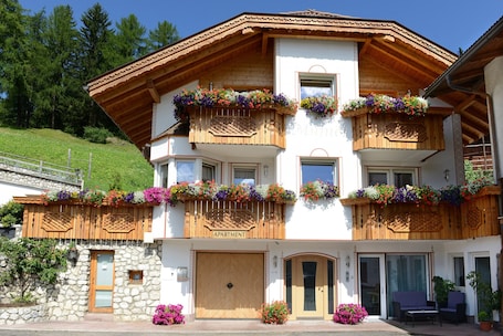 A two-story building with wooden balconies adorned with colorful flower boxes, a stone facade, and a sign indicating it is an apartment.