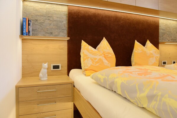 A bedroom with a wooden nightstand, a white cat figurine, a bed with a patterned bedspread, and a stone wall behind the headboard.