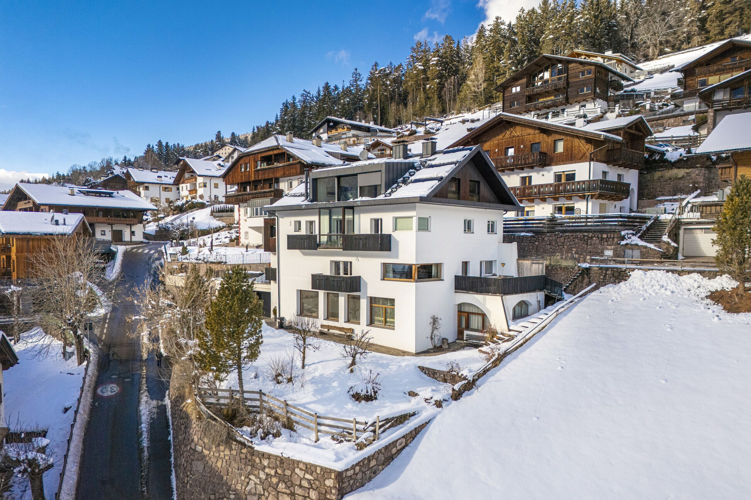 A snow-covered village with traditional and modern buildings, a stone wall, and a clear sky.