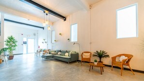 Un hall d’entrée spacieux, avec un grand canapé d’angle gris, des fauteuils en osier et des plantes en pot.