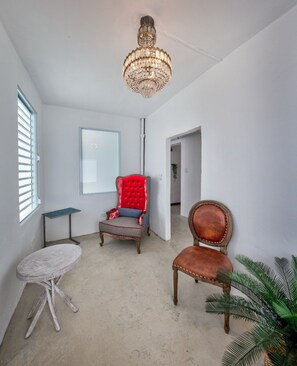 A room with a chandelier, a red upholstered chair, a wooden chair, a small round table, and a potted plant.