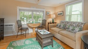 A living room with a beige sofa, a small table, two chairs, a wicker ottoman, a green patterned rug, a window with blinds, a lamp, and a wall-mounted heater. - Rehoboth Beach Efficiency Downtown Deck Sleeps 2 (Rehoboth Beach)