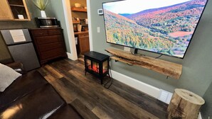 Una sala de estar con una televisión de pantalla plana sobre una estantería de madera, un sofá marrón oscuro y una pequeña mesa con una planta.