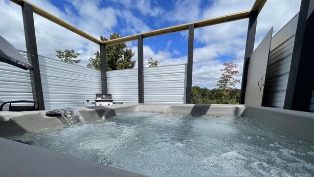 Un jacuzzi con estructura de madera y panel de control, rodeado de paredes metálicas y un cielo despejado.