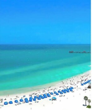 Een strand met wit zand, helder turquoise water en een pier in de verte.