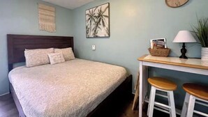 A bedroom with a bed, a desk with two stools, a wall clock, and a plant. - Boho Bungalow! Guesthouse near Downtown & Beaches!  (St. Petersburg)